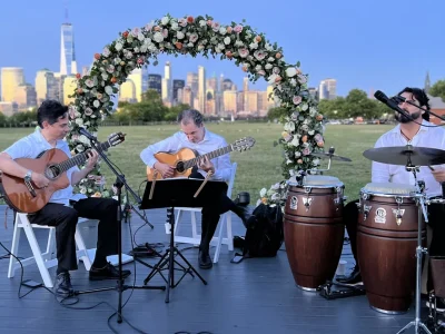 Tomas Rodriguez performing with a guitar and percussion trio at an outdoor event overlooking the New York skyline.